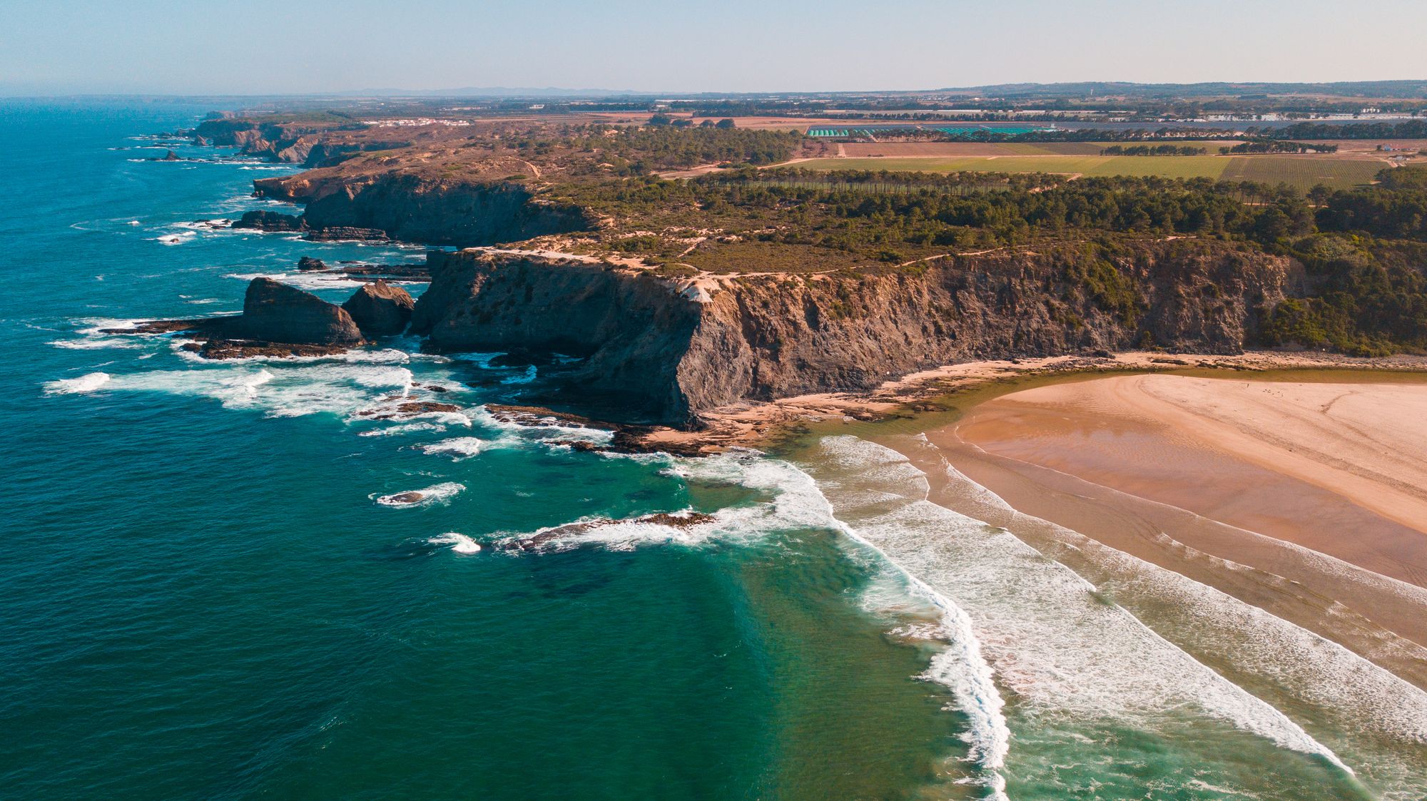 An aerial view of a green coastal headland above a pale beach and turquoise surf